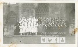 Devant l’église sont regroupés de jeunes filles en blanc et les jeunes garçons en costume de premier communiant entourés par deux prêtres